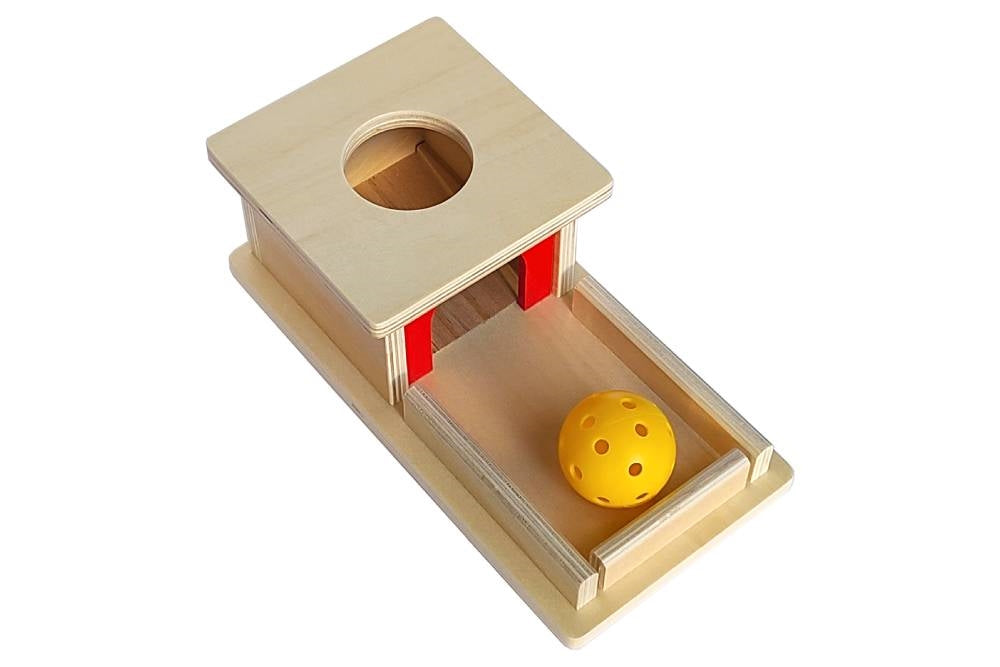 Object permanence box with tray. Natural wood with red accent and yellow hollow plastic ball. Box has a circular hole for the ball to be dropped into, and a ramp for the ball to roll back out on.
