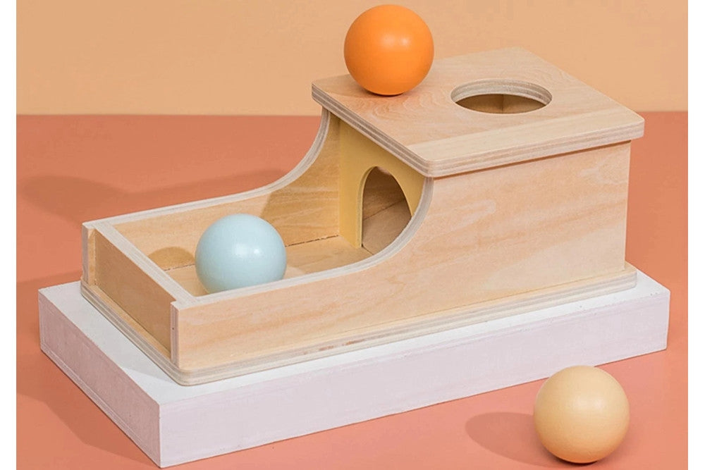 Object permanence box with tray. Natural wood with three balls in light blue, yellow, and orange. Box has a circular hole for the ball to be dropped into, and a ramp for the ball to roll back out on.