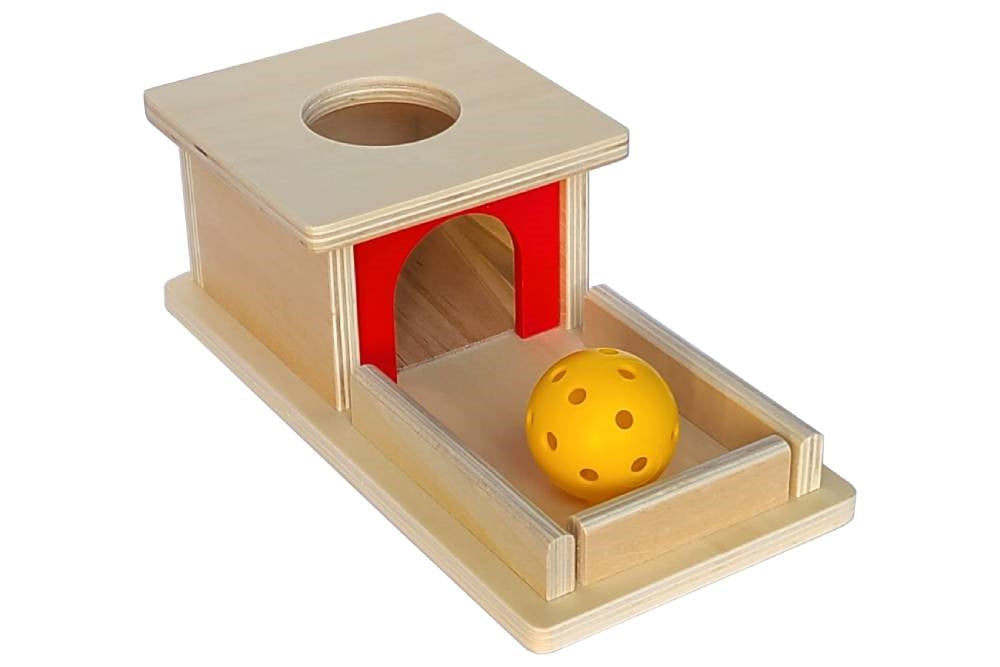 Object permanence box with tray. Natural wood with red accent and yellow hollow plastic ball. Box has a circular hole for the ball to be dropped into, and a ramp for the ball to roll back out on.